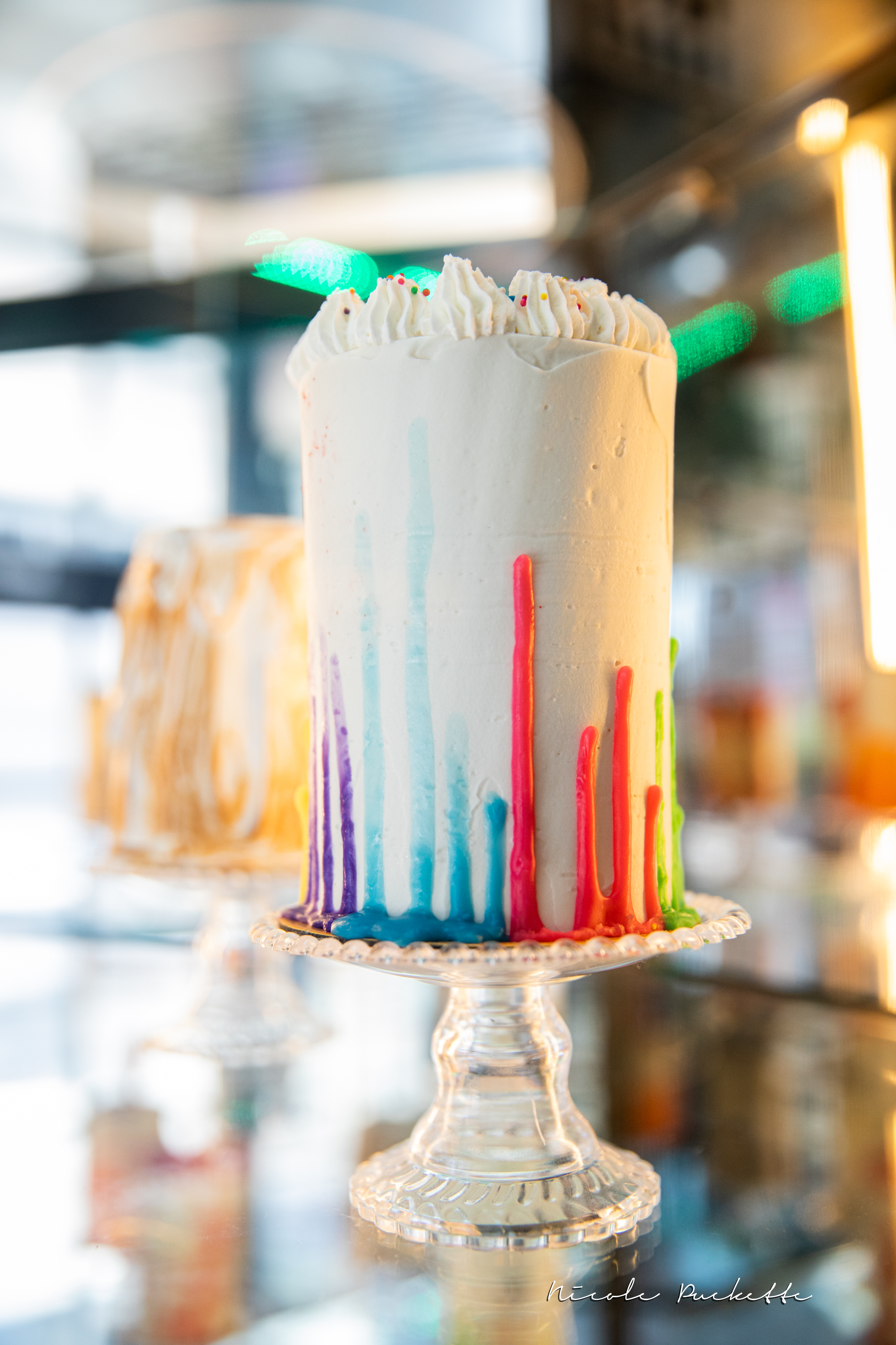 A tall rainbow drip cake on a glass stand at Kakes