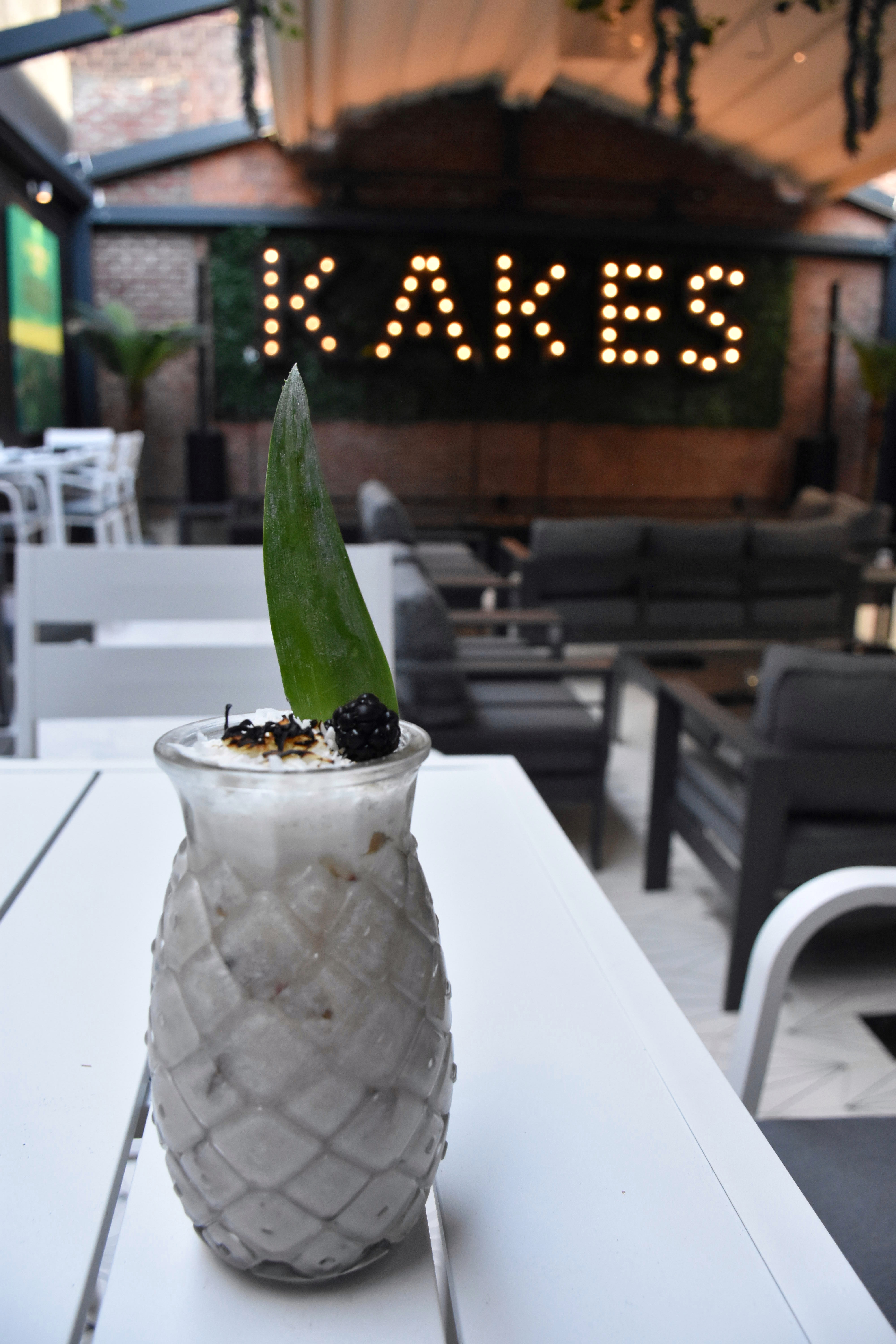 A coconut mocktail in a pineapple glass on a Garden table, the lit KAKES marquee glowing behind it